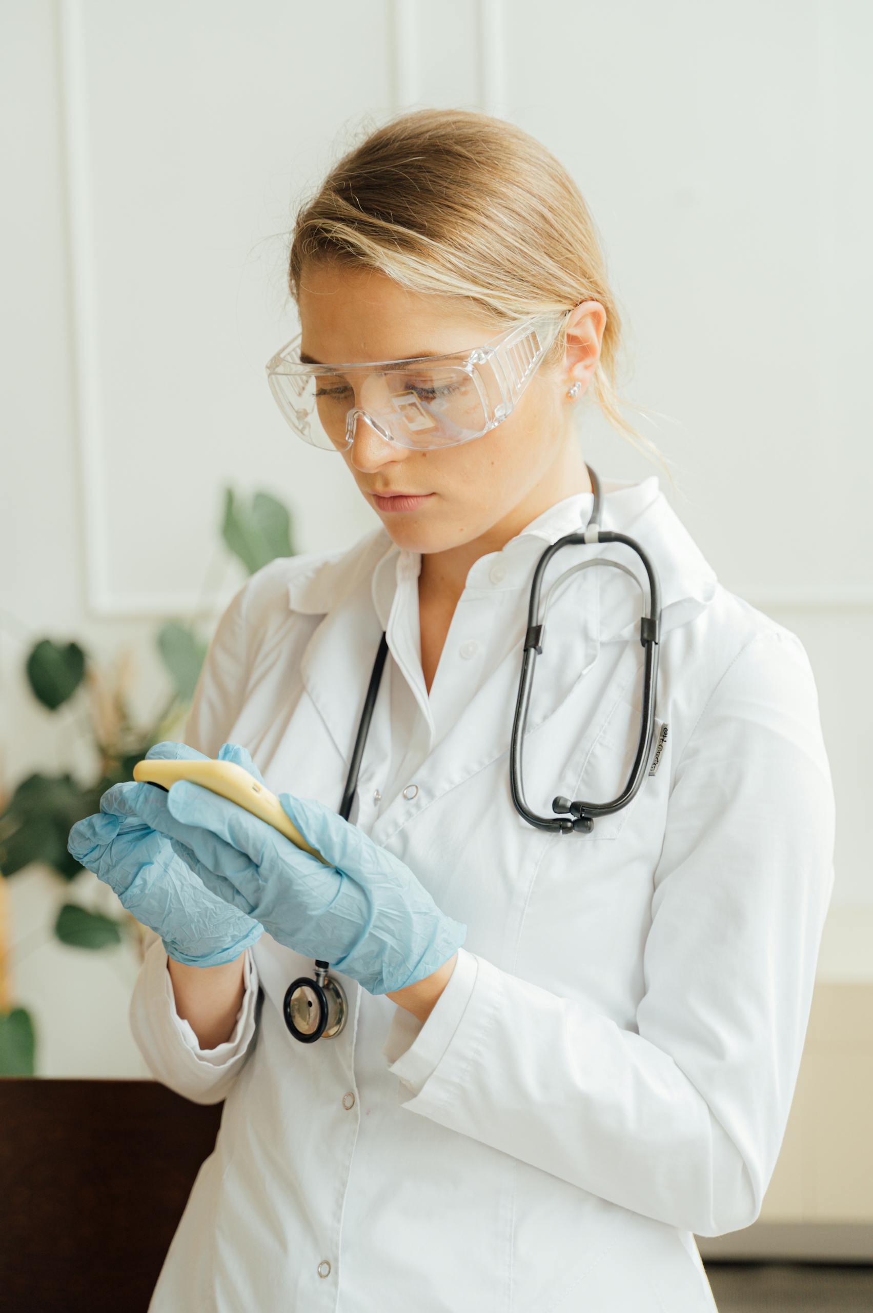 A female doctor with a stethoscope and protective eyewear using a smartphone inside a medical facility.