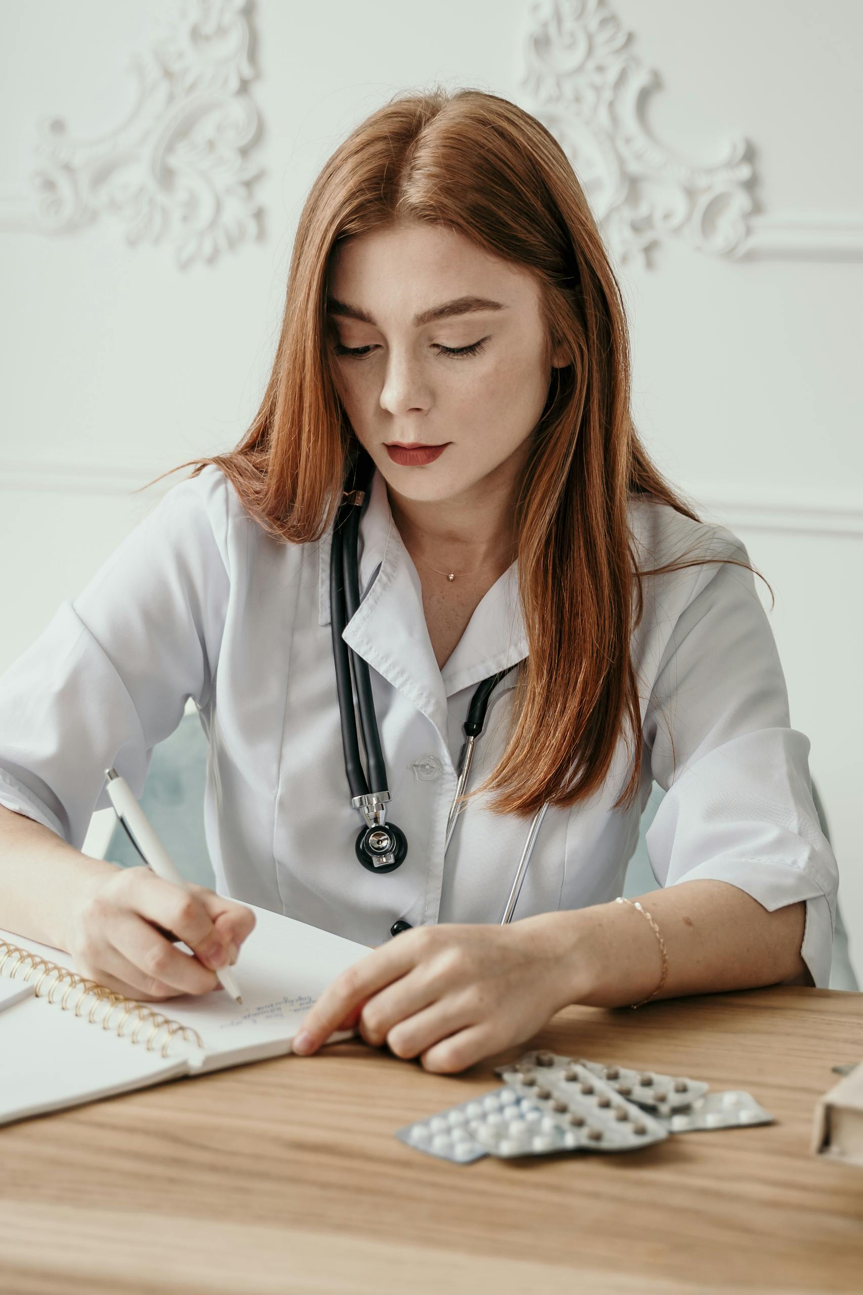 Young female doctor writing notes at her desk with stethoscope and medication in view.
